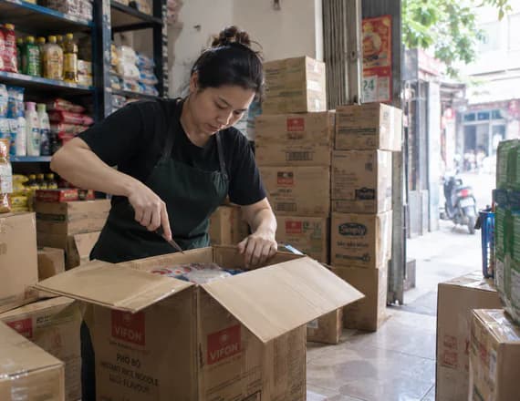 A shopkeeper cutting open a delivery among stacked cartons